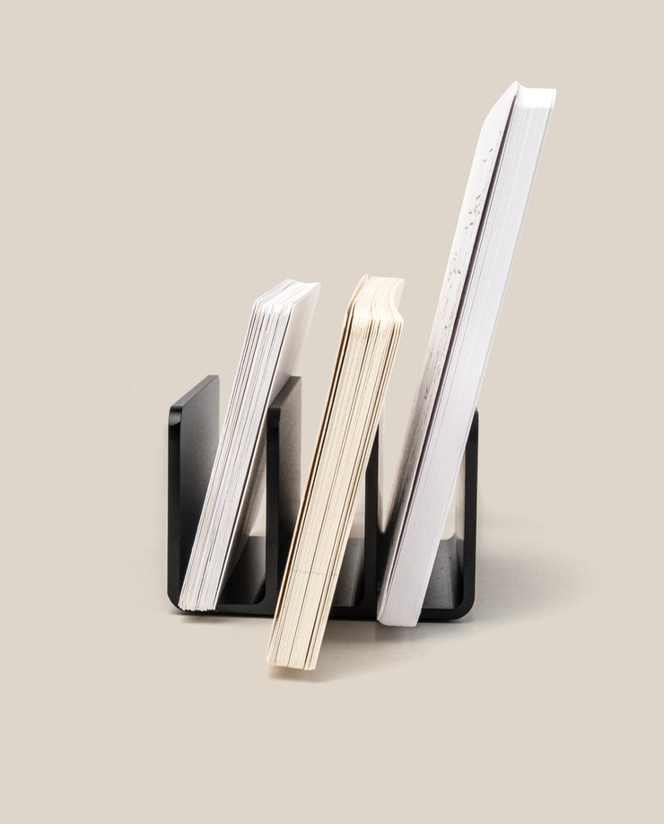 The Analog Card Organizer (Black Aluminum) holds three notebooks of varying sizes upright against a neutral beige background, arranged from smallest to largest with their covers and pages visible side-on.