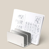 The Analog Card Organizer (Silver Aluminum) has a sleek, minimalist design that doubles as a stand, propping up an open notebook with dotted pages filled with sketches and notes, all set against a plain beige background.