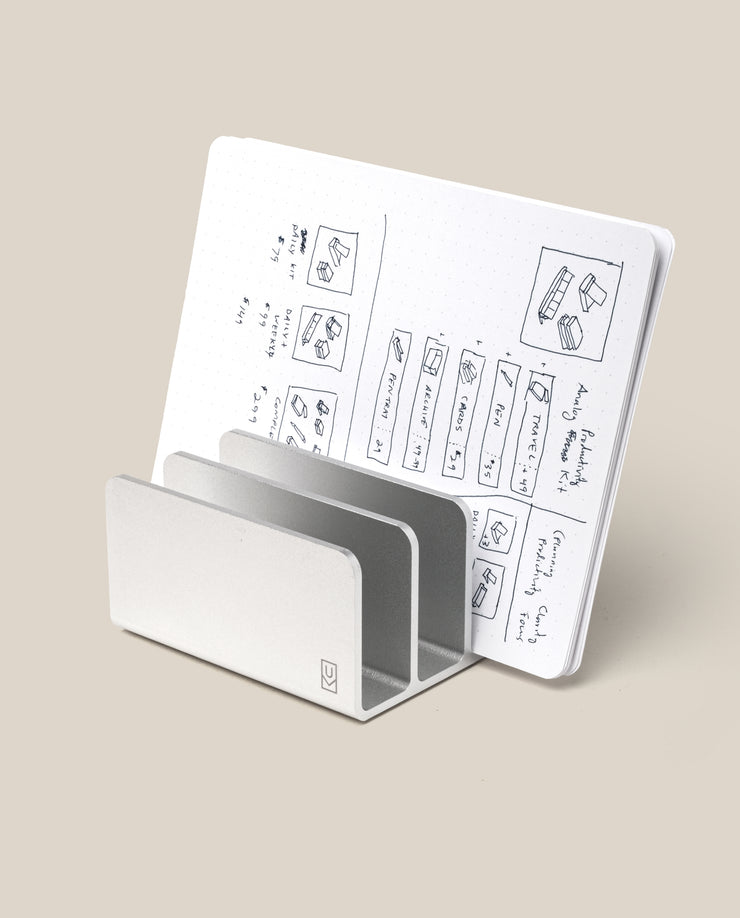 The Analog Card Organizer (Silver Aluminum) has a sleek, minimalist design that doubles as a stand, propping up an open notebook with dotted pages filled with sketches and notes, all set against a plain beige background.