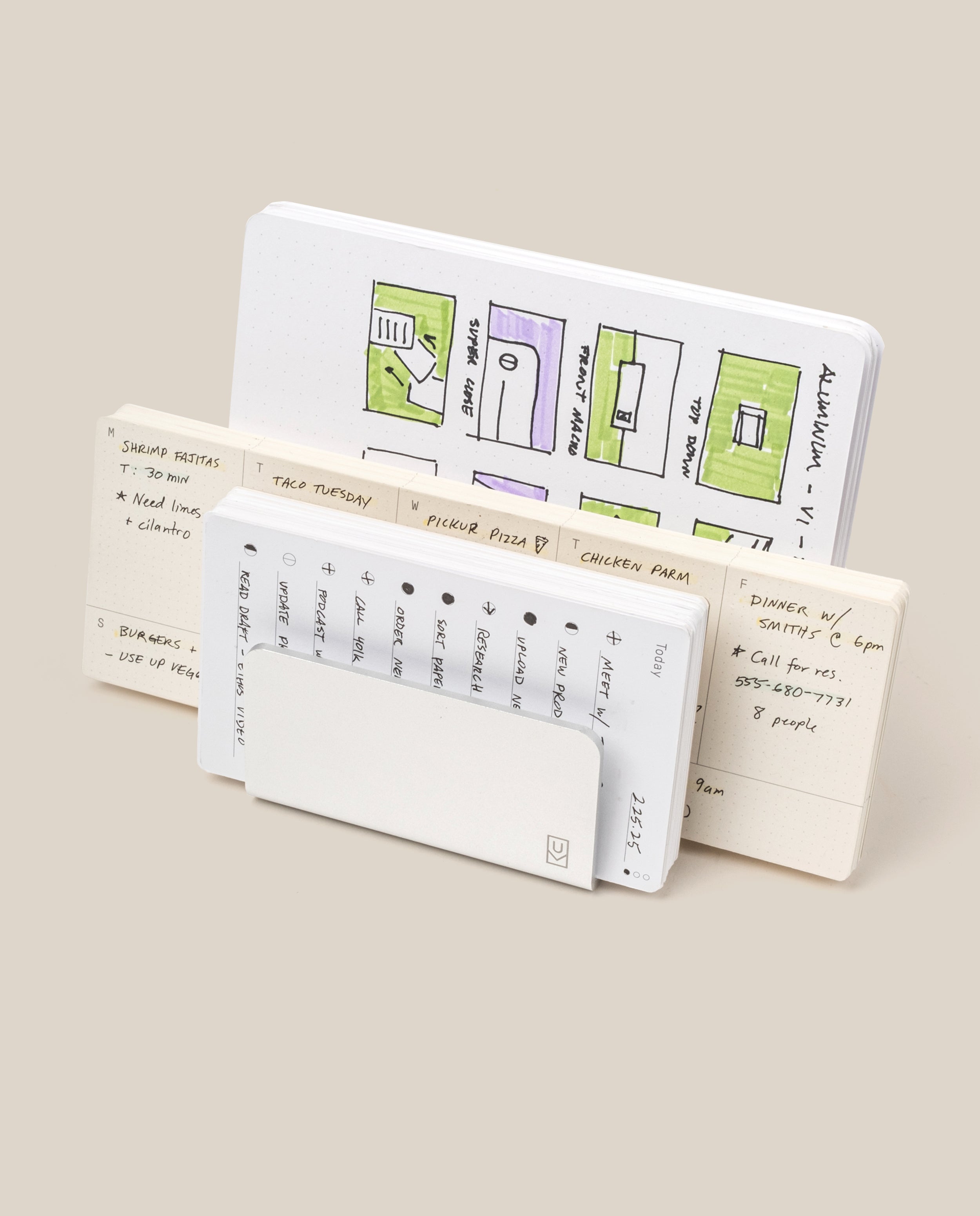 Three open notebooks stand upright. The back features colorful floor plans; the middle, handwritten notes and a meal plan. In front, a small calendar peeks from its cover next to the Analog Card Organizer (Silver Aluminum) for sleek card storage.