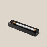 A white and gold pen engraved with "UGMONK" rests on the Analog Pen Tray (Black Aluminum), serving as a sleek pen holder and stylish desk organizer against a beige background.