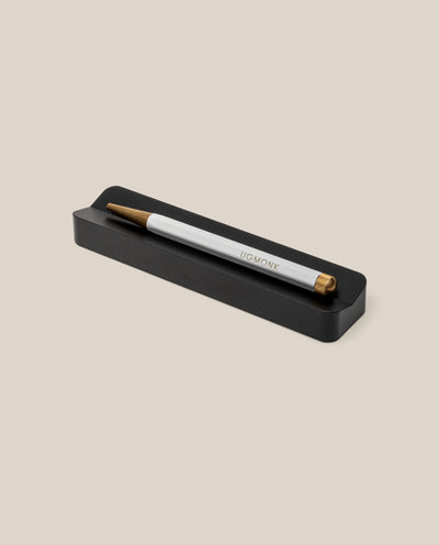 A white and gold pen engraved with "UGMONK" rests on the Analog Pen Tray (Black Aluminum), serving as a sleek pen holder and stylish desk organizer against a beige background.