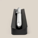A pen from The Complete Analog Collection (Black Aluminum) with a ribbed grip lies horizontally on a black rectangular holder—an elegant desk accessory, shown tip-first against a plain beige background.