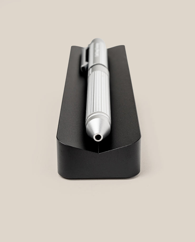 A pen from The Complete Analog Collection (Black Aluminum) with a ribbed grip lies horizontally on a black rectangular holder—an elegant desk accessory, shown tip-first against a plain beige background.