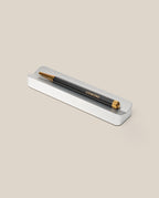A black and gold pen with UGMONK inscribed on its side rests in the Analog Pen Tray (Silver Aluminum), creating an elegant desk accessory against a beige background.