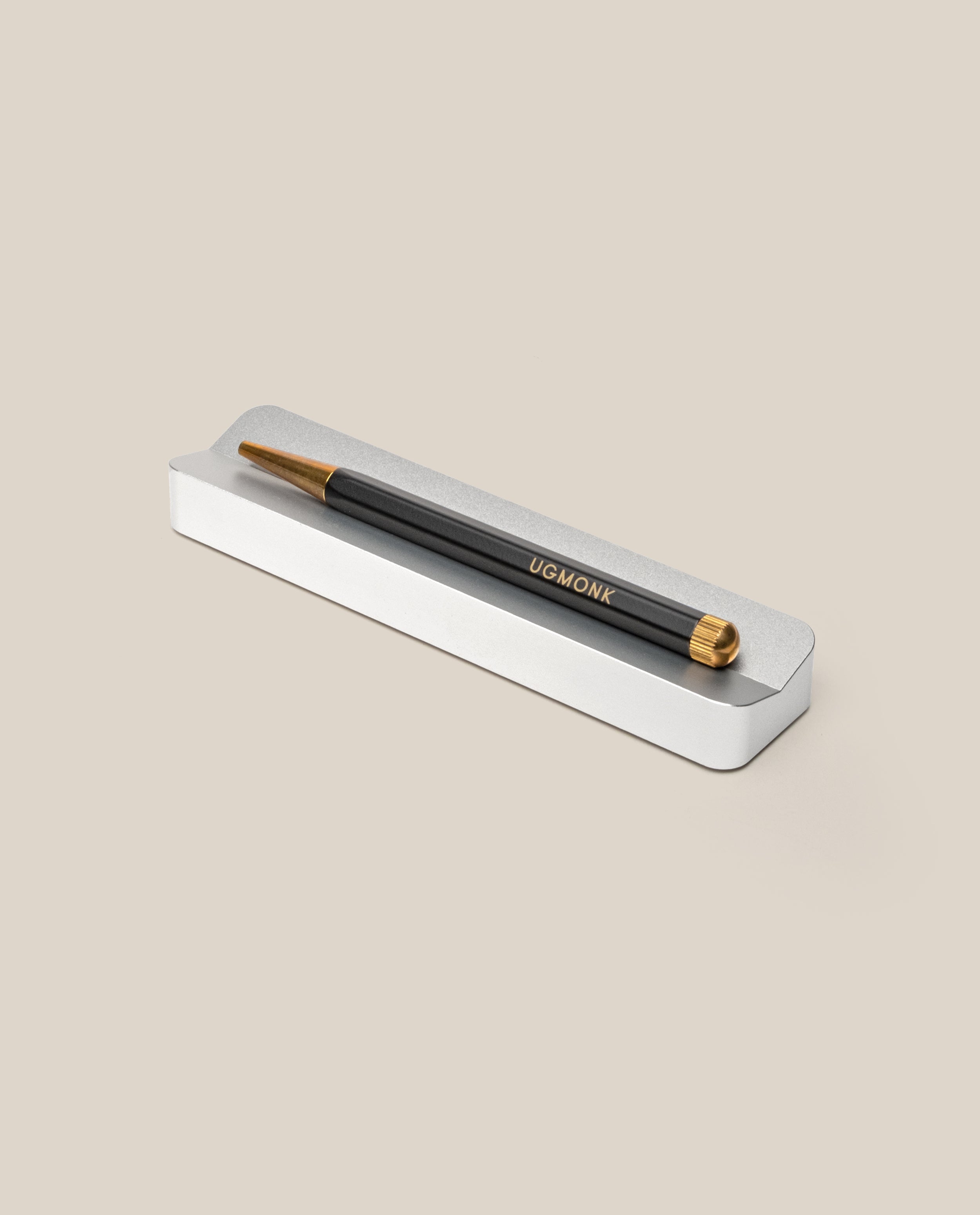 A black and gold pen with UGMONK inscribed on its side rests in the Analog Pen Tray (Silver Aluminum), creating an elegant desk accessory against a beige background.