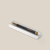 A sleek black and gold UGMONK pen rests in the minimalist Analog Pen Tray (Silver Aluminum) on a beige background—an elegant desk accessory.