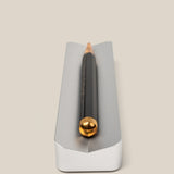 A black pencil with a gold-colored end lies horizontally in The Complete Analog Collection (Silver Aluminum)—a sleek, silver rectangular desktop organizer with a groove—on a beige background.