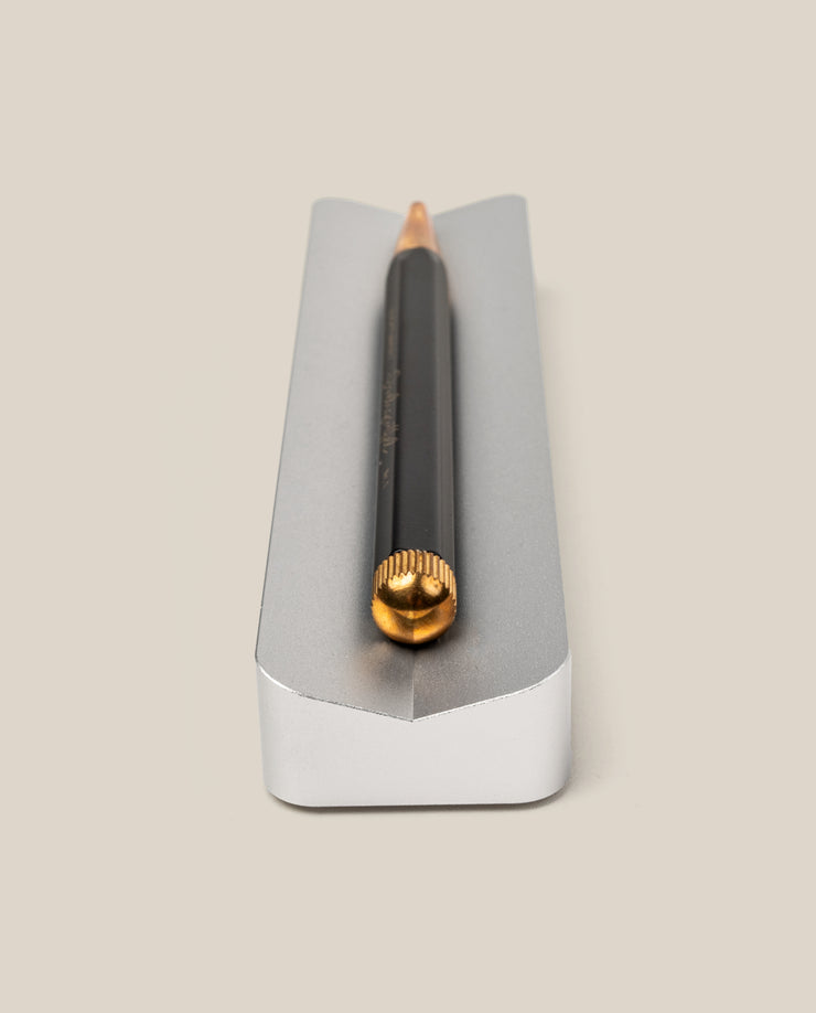 A black pencil with a gold-colored end lies horizontally in The Complete Analog Collection (Silver Aluminum)—a sleek, silver rectangular desktop organizer with a groove—on a beige background.