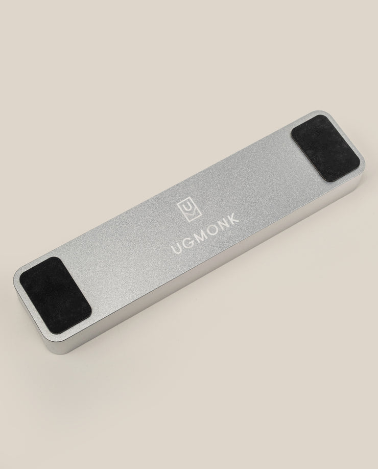 The Analog Pen Tray (Silver Aluminum) is a solid aluminum rectangular pen tray with UGMONK branding and logo on top, two black rectangular pads on each end, displayed on a light beige background—a sleek desk accessory.