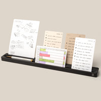 The Analog Card Bar (Large 16" – Black), a solid aluminum desk organizer tray, holds a pencil and four note cards with sketches and notes on a light beige background. Text at bottom: “*Cards sold separately.”.