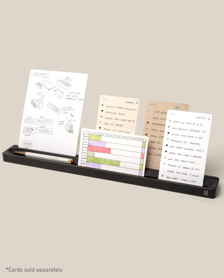 The Analog Card Bar (Large 16" – Black), a solid aluminum desk organizer tray, holds a pencil and four note cards with sketches and notes on a light beige background. Text at bottom: “*Cards sold separately.”.