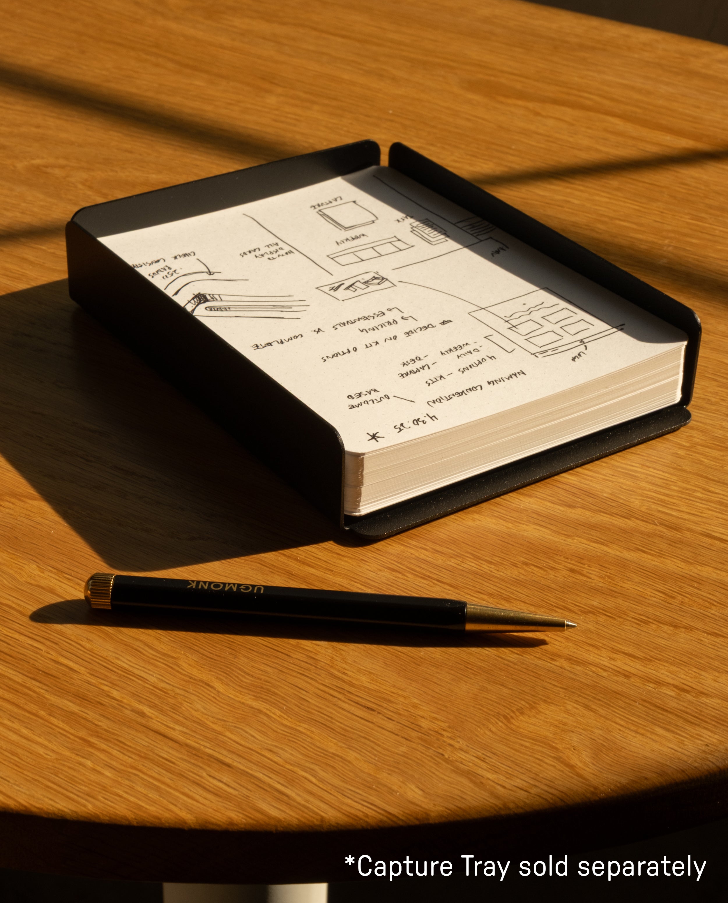 A black pen and notebook with sketches and notes rest on a round wooden table in sunlight. Text in the corner reads, *Capture Tray sold separately. Perfect for jotting ideas or pairing with Analog Dot Grid Cards - Large (12-Pack).