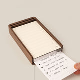 A hand slides a completed task card under a stack of blanks in the wooden tray from the Analog Daily Focus Kit + 3 Refills (Walnut). Text on image: Slide completed cards under the stack—a simple analog productivity system.