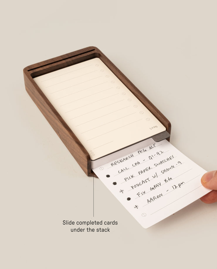 A hand slides a completed task card under a stack of blanks in the wooden tray from the Analog Daily Focus Kit + 3 Refills (Walnut). Text on image: Slide completed cards under the stack—a simple analog productivity system.