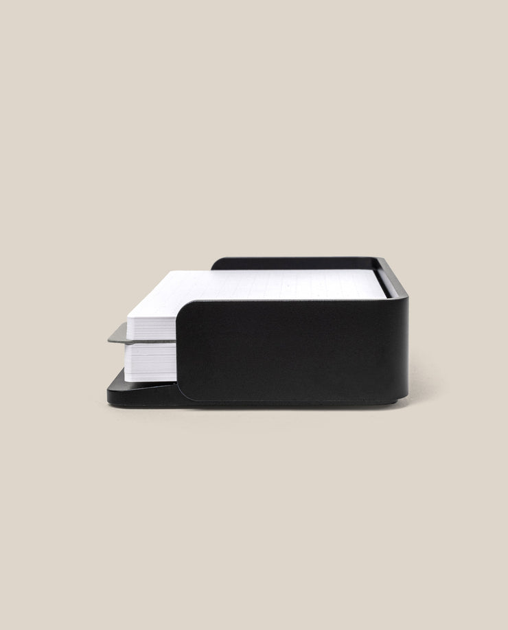 A black Analog Capture Kit - Small (Black Aluminum) holds a stack of white, lined index cards against a plain beige background—ideal for organizing your notes.