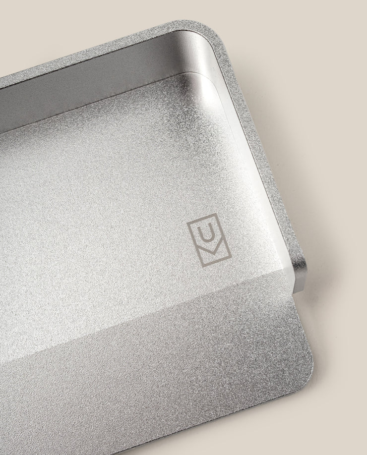 A close-up of the Analog Capture Kit - Small (Silver Aluminum), an aluminum card tray with rounded edges and a small engraved geometric logo, ideal for organizing Small Dot Grid Cards, shown on a beige background.