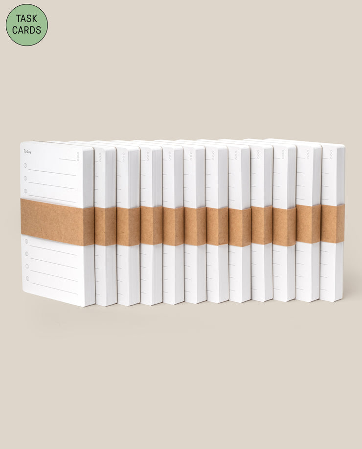 A diagonal row of Analog - Task Cards (12-Pack) notepads, each stacked and wrapped with brown paper bands, is displayed on a beige background. A green circle in the top left highlights them as ideal for analog productivity system fans.