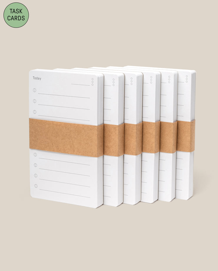 Six stacks of white Analog - Task Cards, each with numbered lines and wrapped in a brown band, are displayed on a light beige background. A green label in the corner reads “TASK CARDS”—ideal for your productivity system.