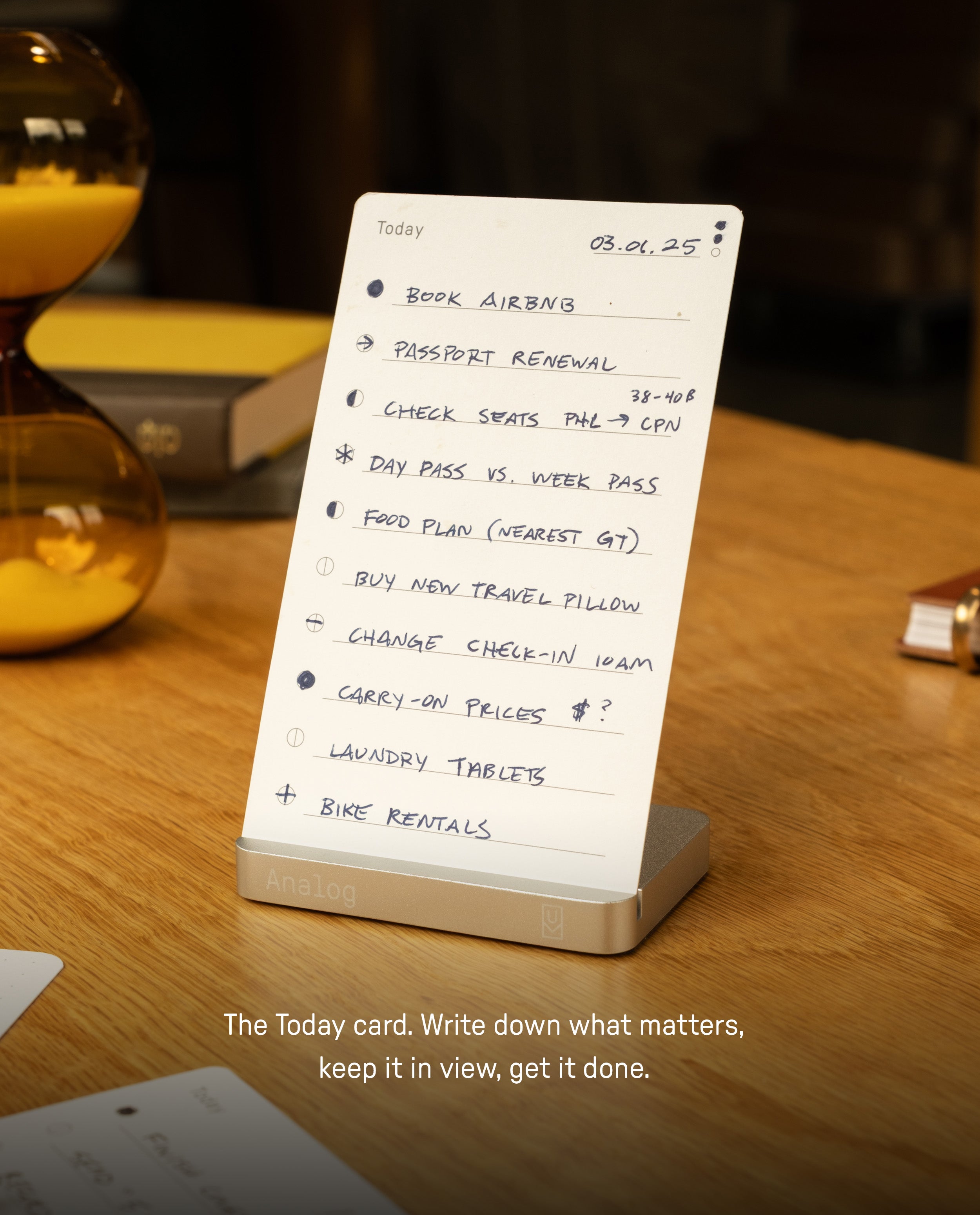 A to-do list featuring tasks like booking Airbnb, renewing a passport, and arranging bike rentals is neatly displayed on a wooden desk with an hourglass nearby. The tasks are organized in stylish visual cards within the Analog Starter Kit (Silver), housed in its sleek aluminum card stand. Text below reads: The Today card. Write down what matters, keep it in view, get it done—an elegant paper card system for staying focused with visual task cards.