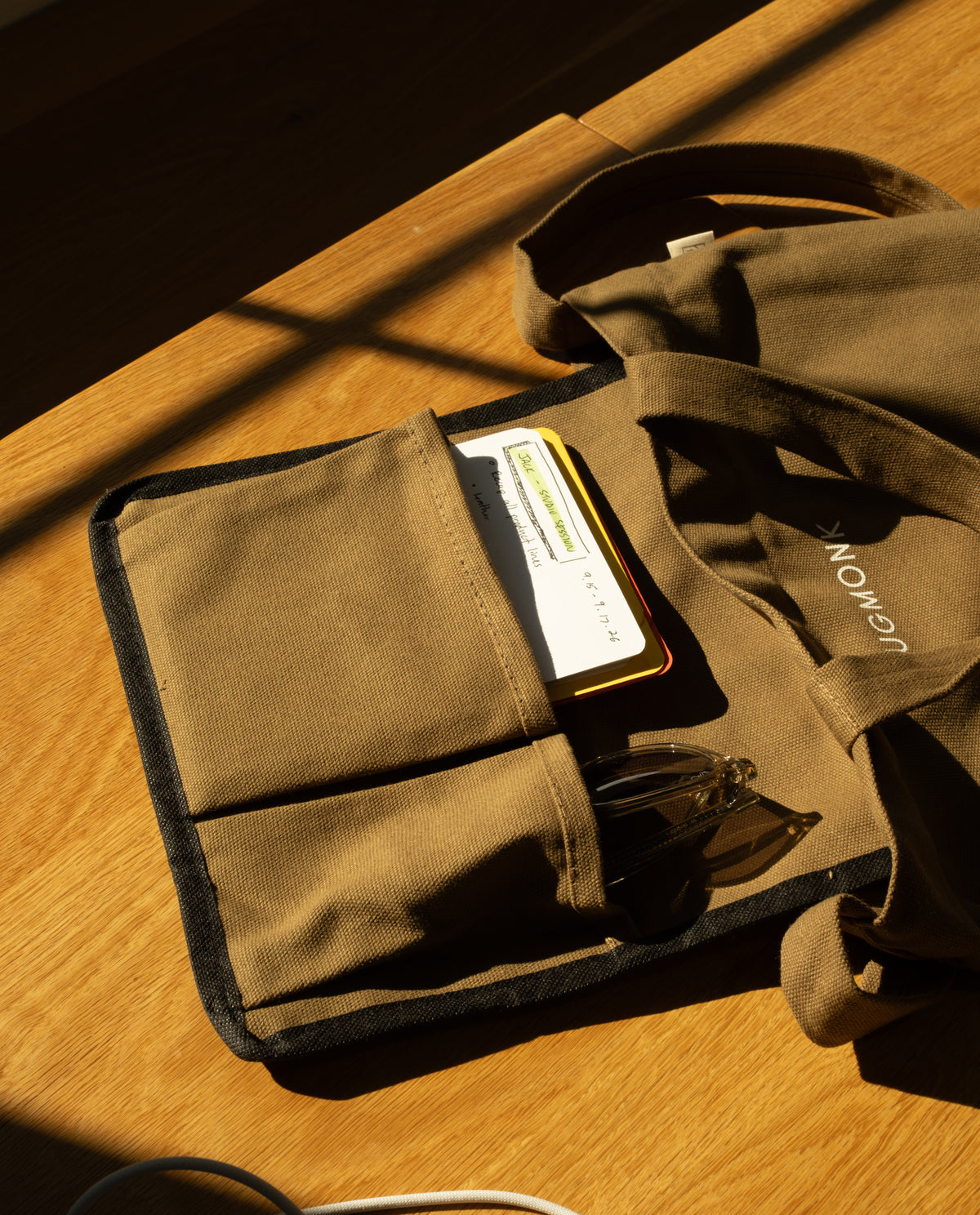A brown heavyweight cotton Utility Tote Bundle - Set of 3 with an open exterior pocket holding a notebook and glasses.