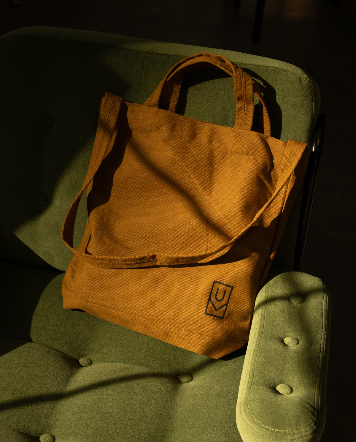 Mustard yellow heavyweight cotton Utility Tote Bundle - Set of 3 with a small black logo on a green armchair.