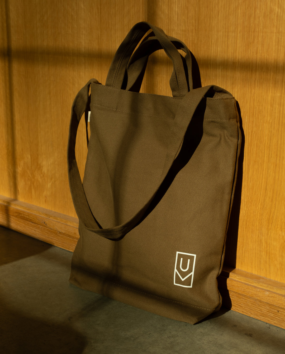 Durable olive green Utility Tote Bundle - Set of 3 canvas tote bag with shoulder straps and a white logo on the lower corner, leaning against a wooden wall.