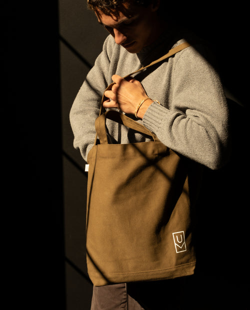 A person wearing a grey sweater holds a Utility Tote Bundle - Set of 3, a durable, brown canvas tote bag with a white logo on the bottom corner.