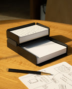 The black steel Capture Bundle (Small Tray + Large Tray) holds two stacks of notecards—one handwritten, one blank—on a wooden table, with a pencil and a sheet of sketches and writing nearby.