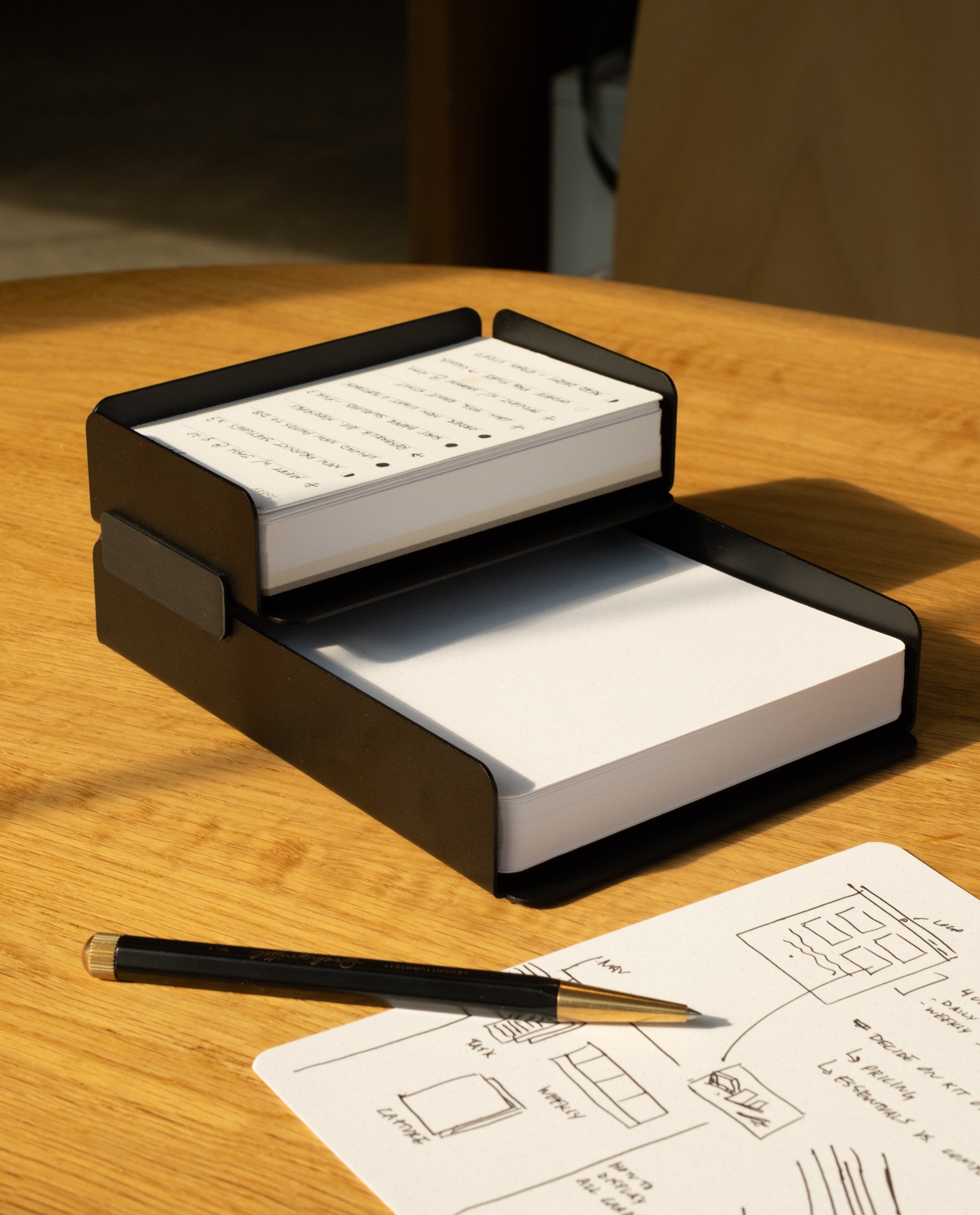 The black steel Capture Bundle (Small Tray + Large Tray) holds two stacks of notecards—one handwritten, one blank—on a wooden table, with a pencil and a sheet of sketches and writing nearby.
