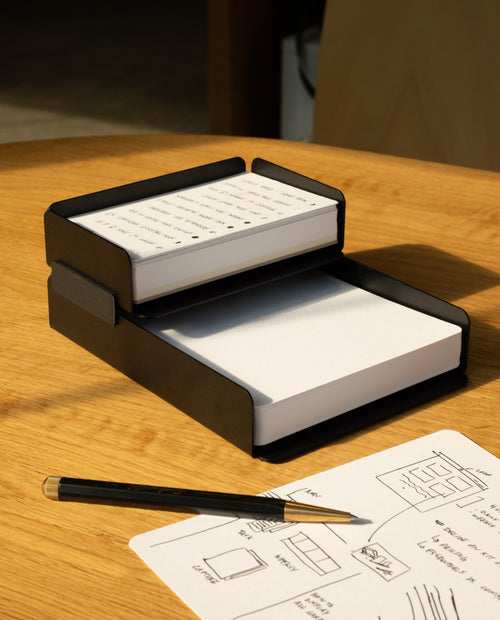 The black steel Capture Bundle (Small Tray + Large Tray) holds two stacks of notecards—one handwritten, one blank—on a wooden table, with a pencil and a sheet of sketches and writing nearby.