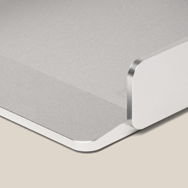 Close-up of a metallic corner of a flat surface, possibly a minimalist desk organizer or laptop stand, on a beige background. The object has a smooth, matte finish and curved edges.