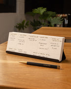 An Analog Weekly Kit (Black Steel) sits on a wooden table with handwritten plans, paired with a black and gold UGMonk pen, a sleek steel card holder, and a leafy plant. Blurred weekly planner cards appear in the background.
