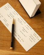 A pen lies on a rectangular notepad with a handwritten schedule, as an open Analog Weekly Kit (Black Steel) sits nearby on the wooden surface, ready to store weekly planner cards.