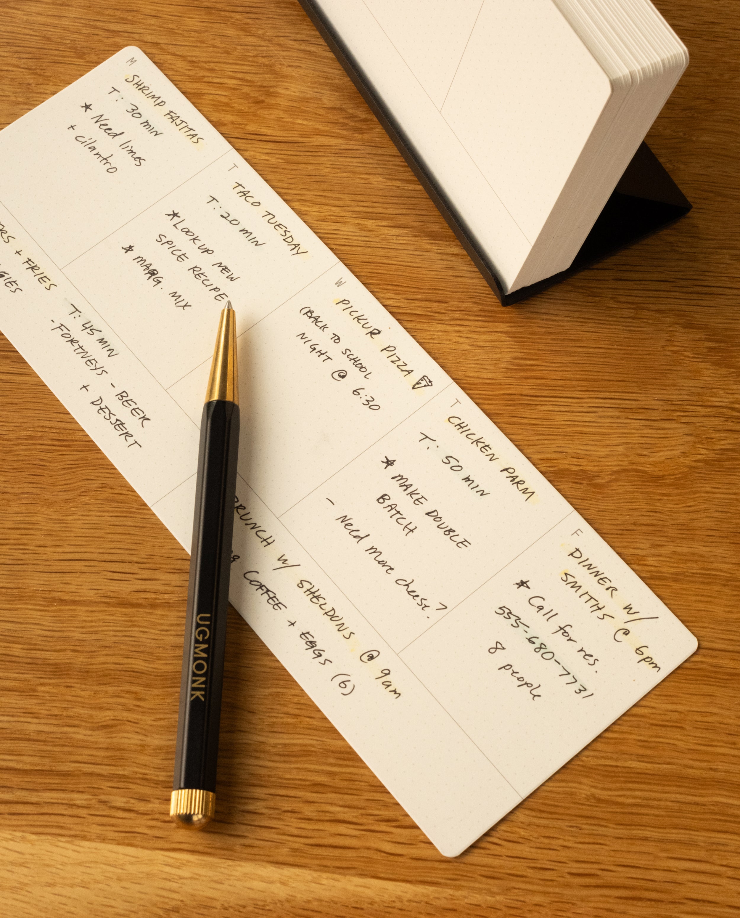 A pen lies on a rectangular notepad with a handwritten schedule, as an open Analog Weekly Kit (Black Steel) sits nearby on the wooden surface, ready to store weekly planner cards.