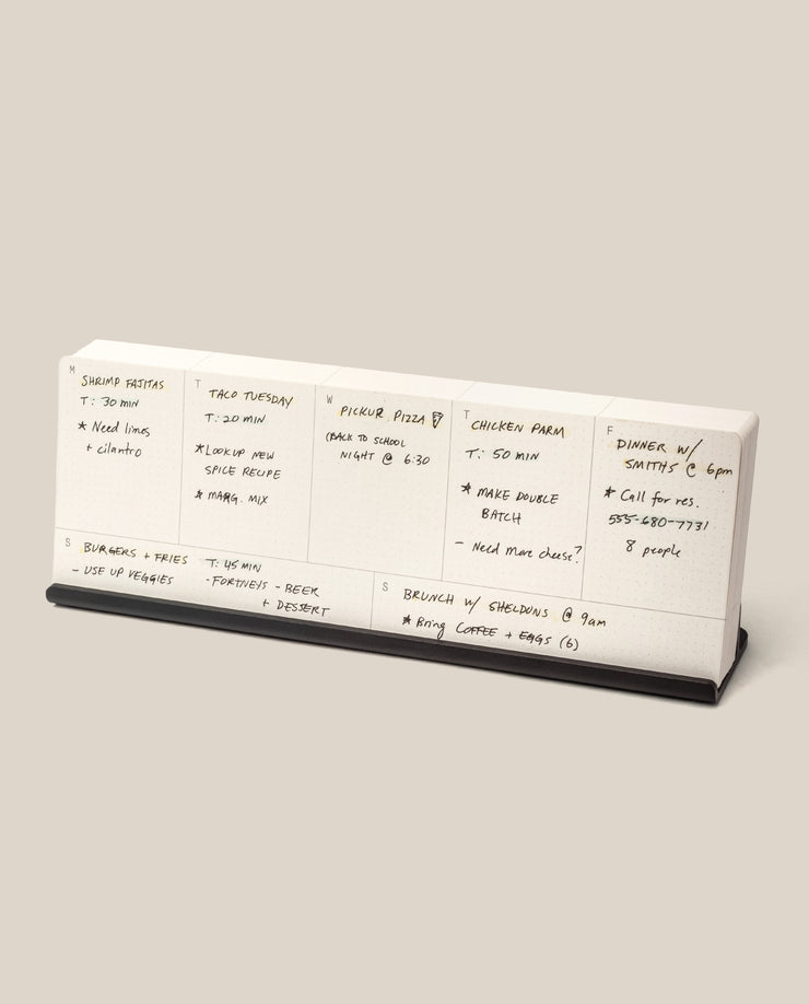 The Analog Weekly Planning Kit (Black Aluminum) is a minimalist desk calendar on a sleek stand, perfect for organizing handwritten meal and event plans like shrimp risotto dinners, takeout nights, and brunch with friends.