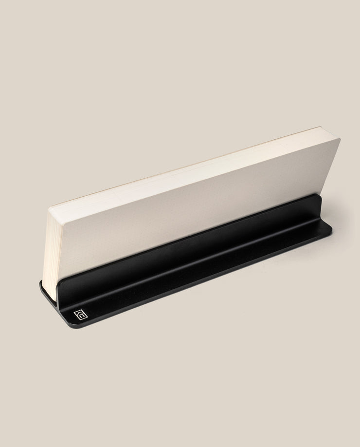 A closed book stands upright in a sleek, black Analog Weekly Planning Kit (Black Aluminum) against a light beige background. The stand supports the bottom edge, ideal for displaying Weekly Cards or organizing your planning kit essentials.