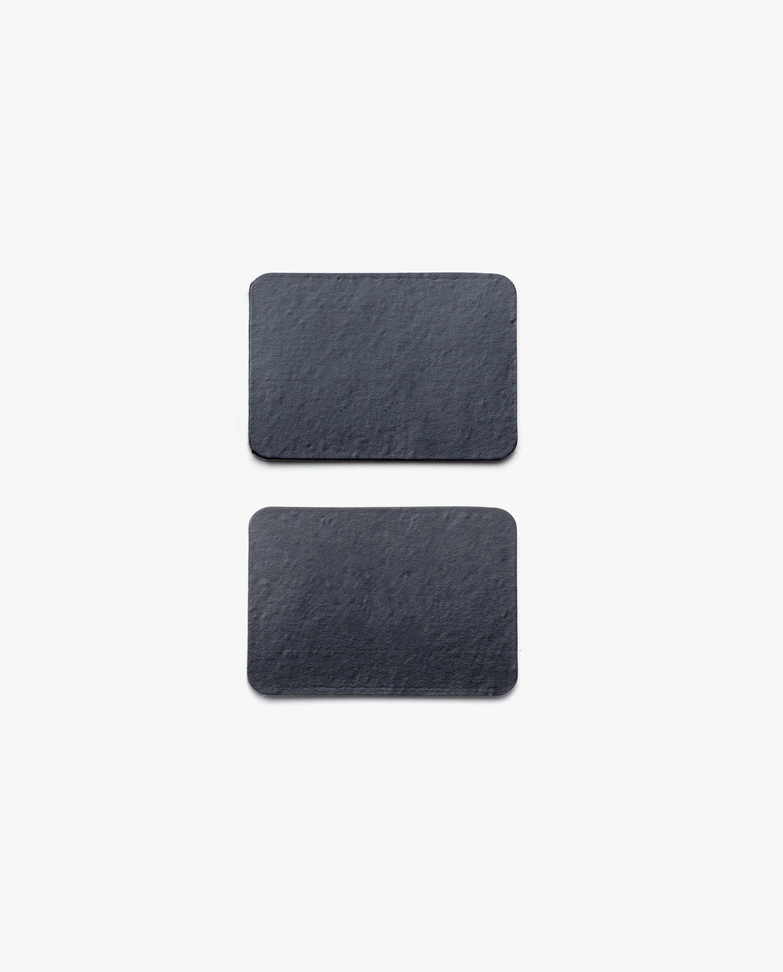 Two rectangular, dark gray slate pieces with rounded corners from the Magnet Kit (Pen Tray and Analog Weekly) are placed vertically on a plain white background, spaced evenly apart—these streamlined accessories are designed to complement your Gather Monitor Stand or Pen Tray. Cleverly integrated magnets help keep these sleek pieces securely in place on your workspace.