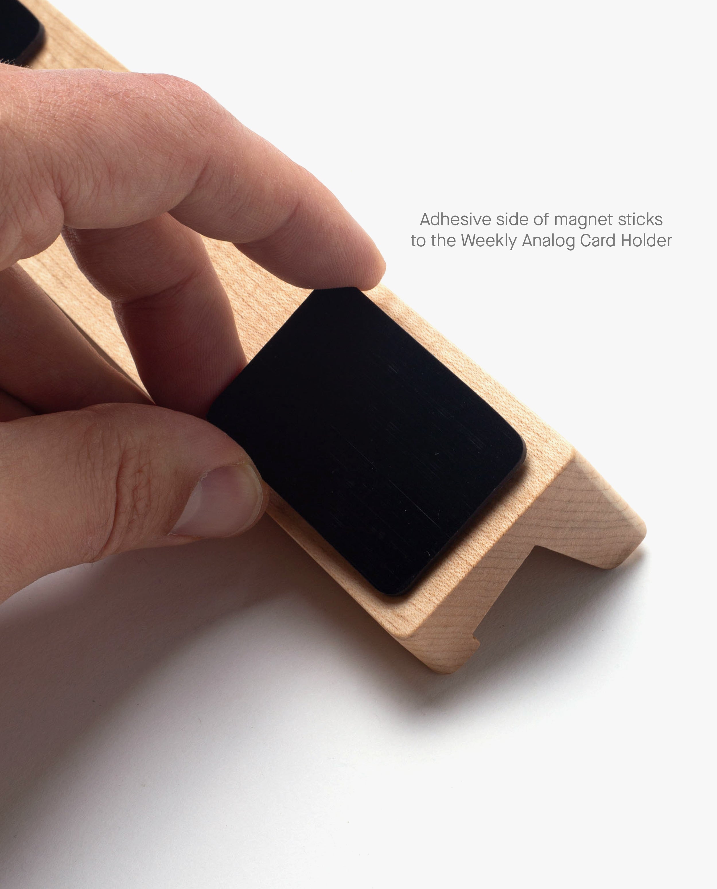 A hand places a black magnet strip with an adhesive side onto a wooden card holder, perfect for organizing your Gather Monitor Stand or Pen Tray. Text reads: The adhesive side of the Magnet Kit (Pen Tray and Analog Weekly) attaches to the Weekly Analog Card Holder.