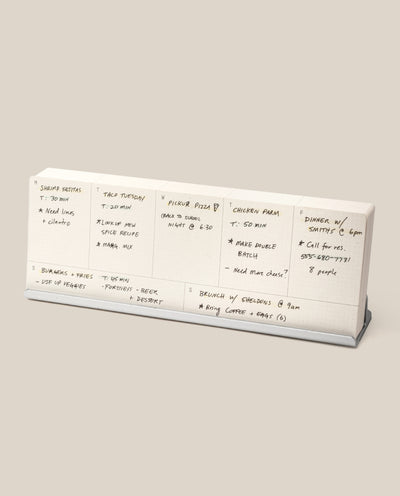 The Analog Weekly Planning Kit (Silver Aluminum) is shown on a stand with handwritten daily notes for meals, prep times, ingredients, and a dinner event with friends. The background is plain beige.