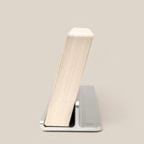 A thick paperback book stands upright, leaning back on The Complete Analog Collection (Silver Aluminum) minimalist book holder against a plain beige background.