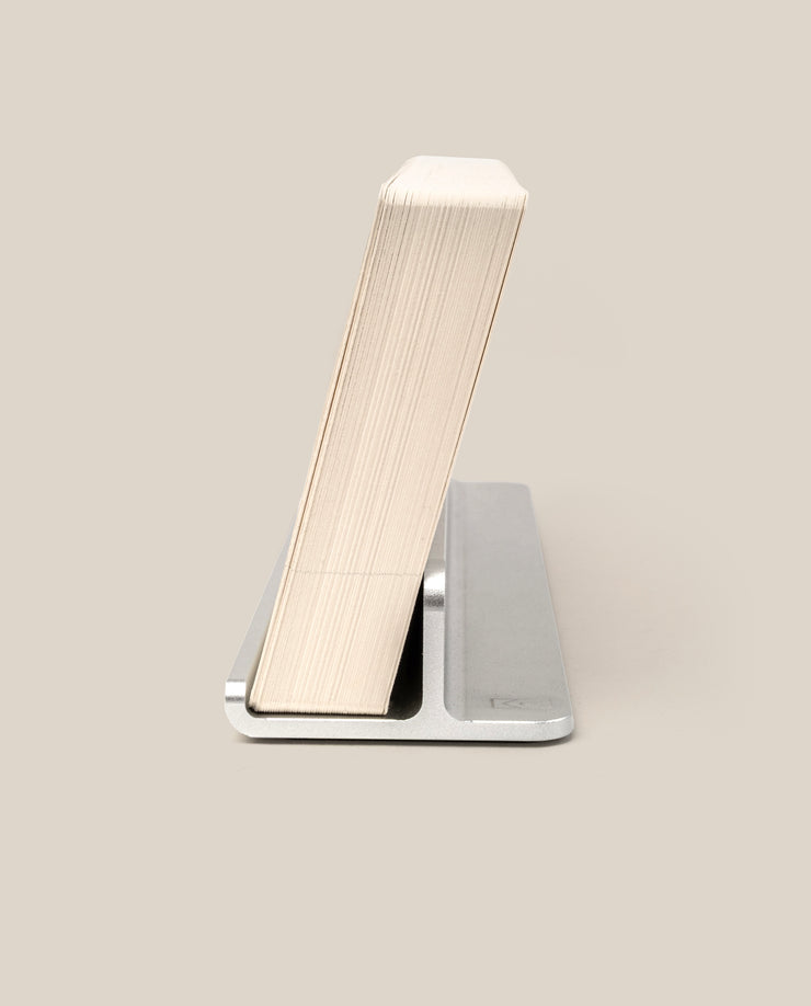 A thick paperback book stands upright, leaning back on The Complete Analog Collection (Silver Aluminum) minimalist book holder against a plain beige background.