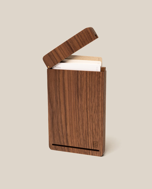 The Analog Travel Case (Walnut) features a smooth walnut finish with a subtly engraved logo on the front bottom corner. Its hinged lid, held securely by a discreet magnetic closure, can be slightly opened to reveal several cards stored inside.