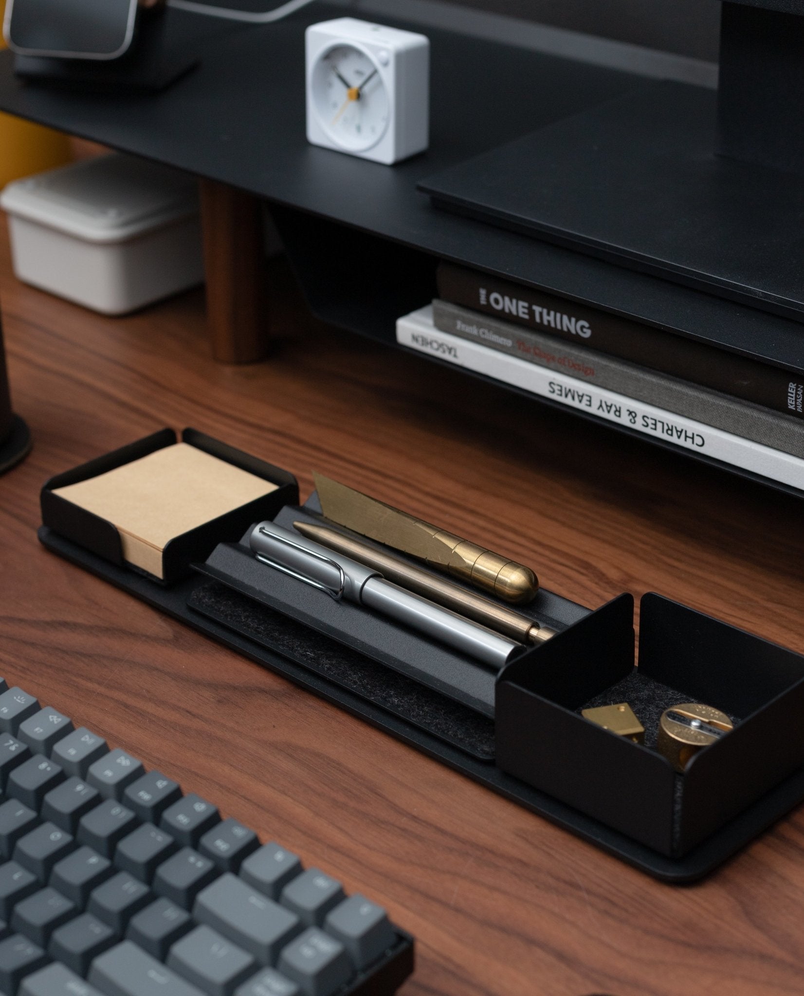 A desk setup featuring a keyboard, pens in a tray, a brass ruler, sticky notes, clips, and magnetic Gather modules on the Gather 1x4 Base Plate (Black), plus stacked books and a small white clock—all on a wooden surface.