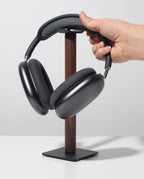 A hand places over-ear black headphones onto the Gather Headphone Stand (1x1 - Black/Walnut), which features a solid walnut wood post and a powder-coated steel base, set on a white surface against a plain light background.