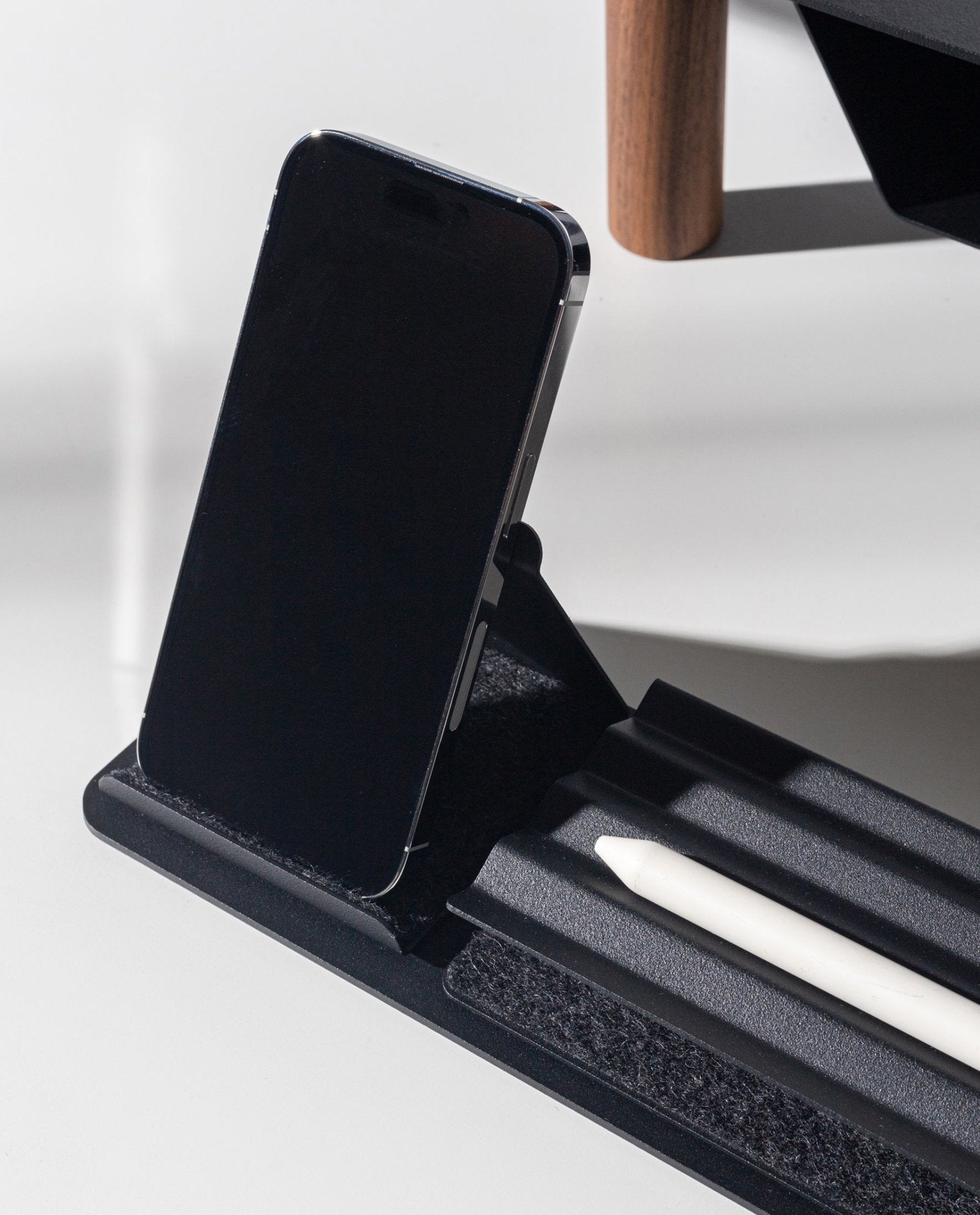 A black smartphone sits upright on the Gather Phone Stand (1x1 - Black) while a white stylus rests in its groove. Sunlight casts clear shadows across the scene.