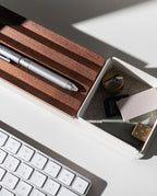 A close-up of a white desk with a keyboard, Gather Small Tray (1x1 - White) as a desk organizer holding a sharpener, eraser, SD card, paper clips, notepads, and wooden pen holder. Sunlight casts shadows across the items. Pen holder is made in Pennsylvania.