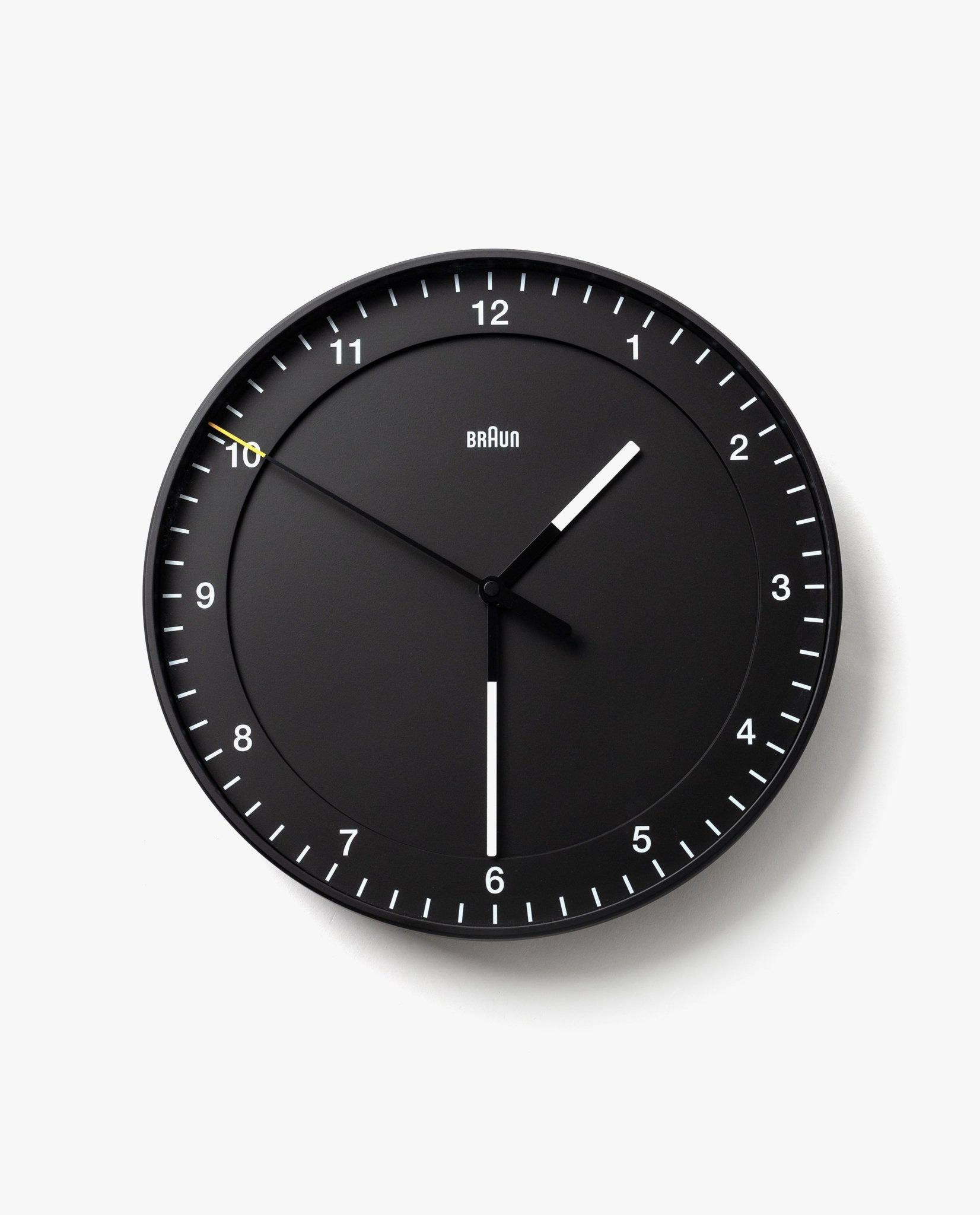 The Braun Wall Clock (Black) features white hour and minute hands, a slim yellow second hand, and silent operation. Clear white tick marks and numbers on a plain white background make the clock easy to read, displaying 10:30:50.