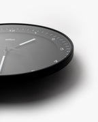 The Braun Wall Clock (Black) features minimalist white hour markers and hands, shown at an angle with only the lower right of the face visible against a white background. An easy-to-read clock, ideal for any modern space.