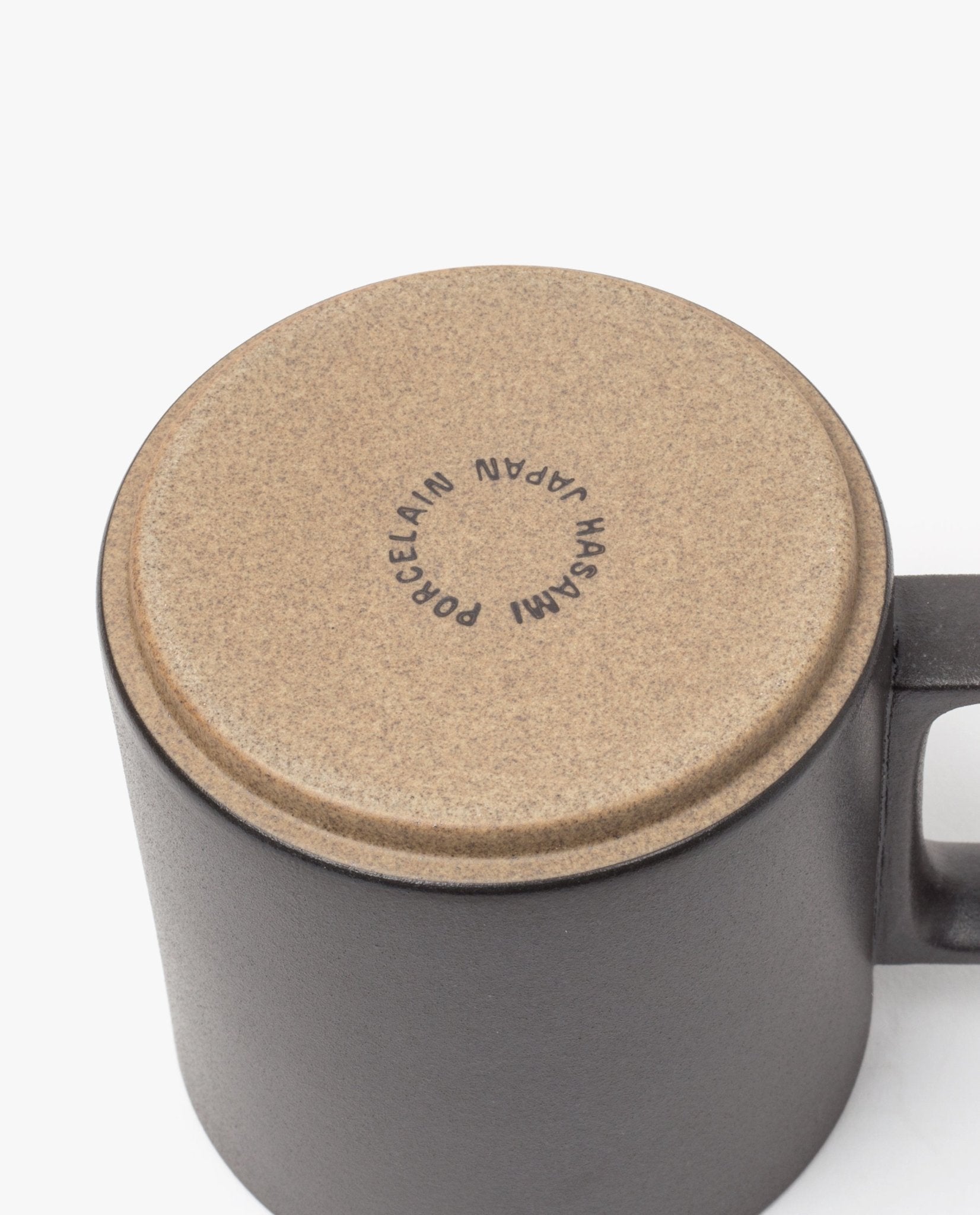 A close-up of an upside-down Hasami Porcelain Mug (Matte Black) shows its tan bottom stamped with "HASAMI PORCELAIN JAPAN," emphasizing its handmade quality. The mug’s handle is visible on the right side.